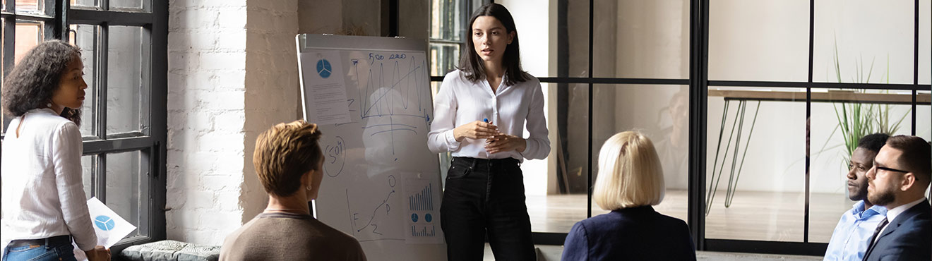 A young professional stands by a whiteboard covered in notes and charts and speaks to a group of other professionals.