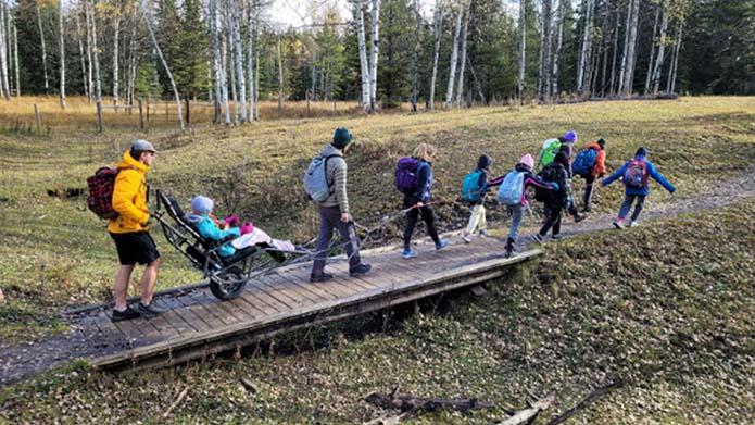 A group of young students walk across a bridge. One student is using the HPED TrailRider and being pushed by an adult.