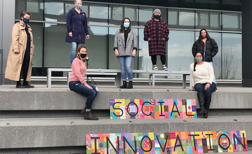 Catherine Pearl, Wendy Lees, Danika Dinko, Kat Bravo, Ashley Villaflores, Roshni McCartney, Ivy Daminan standing in front of a handcrafted sign that reads 'Social Innovation' at the Riddell Library and Learning Centre.