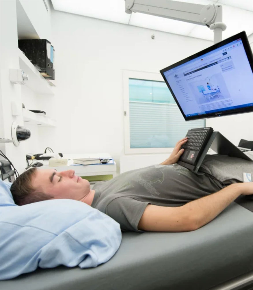 A man lying on a hospital bed looking at a computer monitor.
