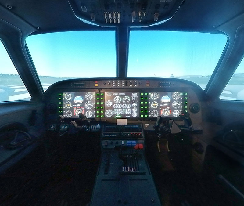 Image of the inside of the aviation simulator. It has windows that look on a screen projected with a runway and mand dials and buttons of a plane.
