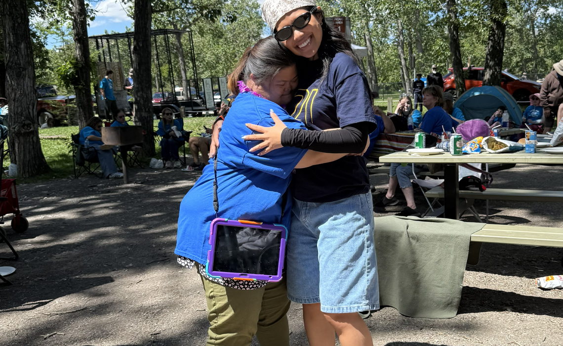 Aisyah Ghazali wearing sunglasses and a bandana, smiling at the camera while sharing a warm hug with a woman wearing a blue shirt and carrying a cross-body tablet at a sunny outdoor picnic.