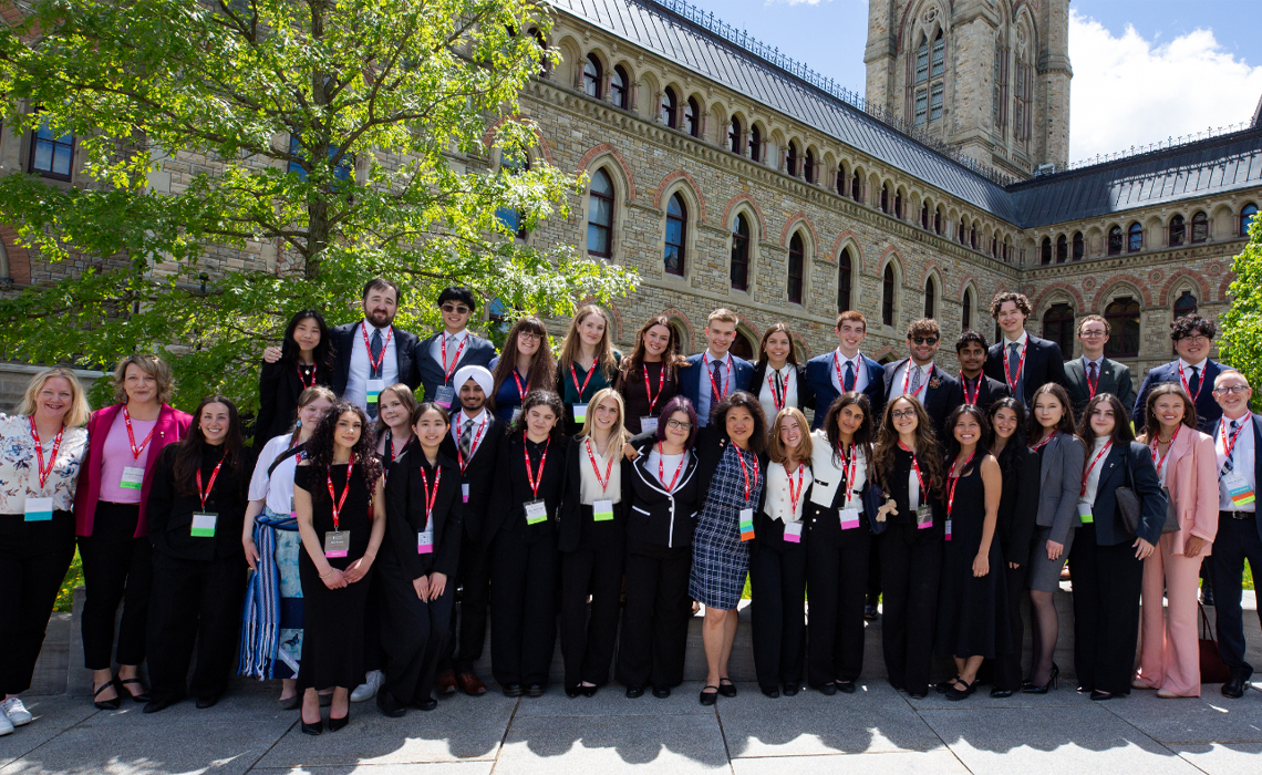 A large group of people in formal business attire and red event lanyards posing together for a group photo outdoors.