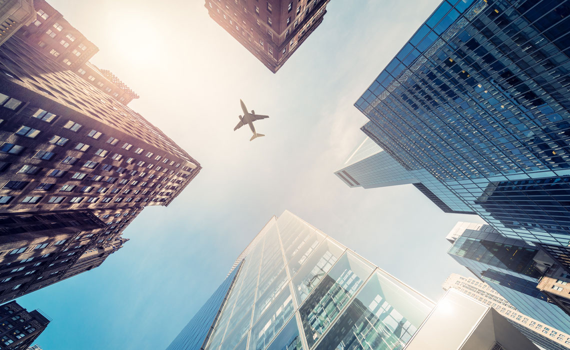 Airplane in flight above cityscape with skyscrapers and urban infrastructure visible.