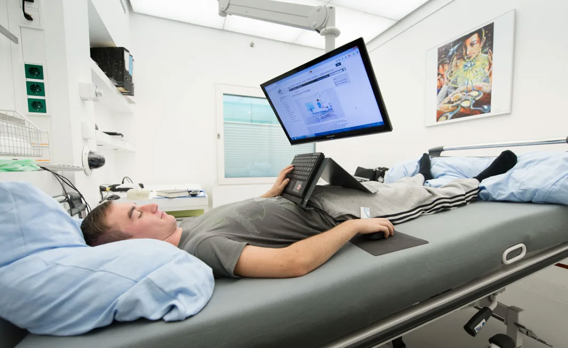 A man lying on a hospital bed looking at a computer monitor.