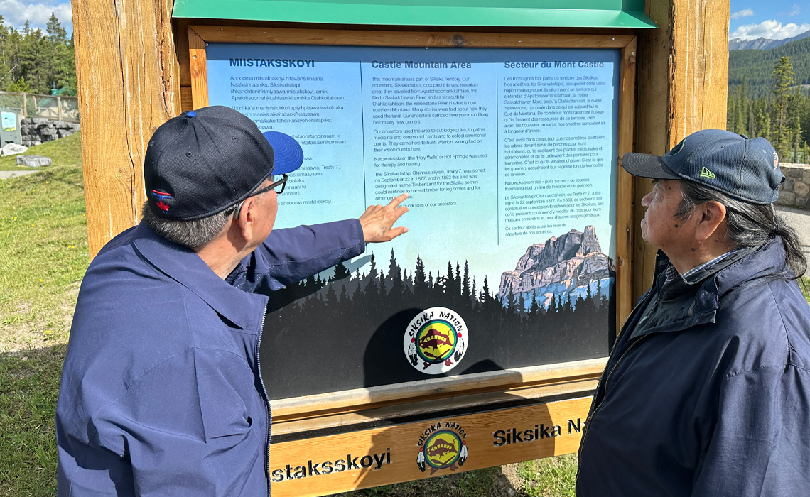 Elders Roy Bear Chief and Hayden Melting Tallow in front of an interpretive signage telling the story of Miistaksskoyi (Castle Mountain).