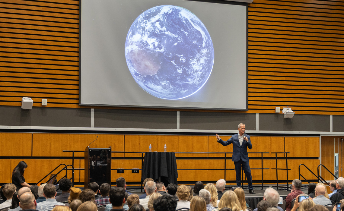Colonel Chris Hadfield presenting to a large audience in a well-lit conference hall.