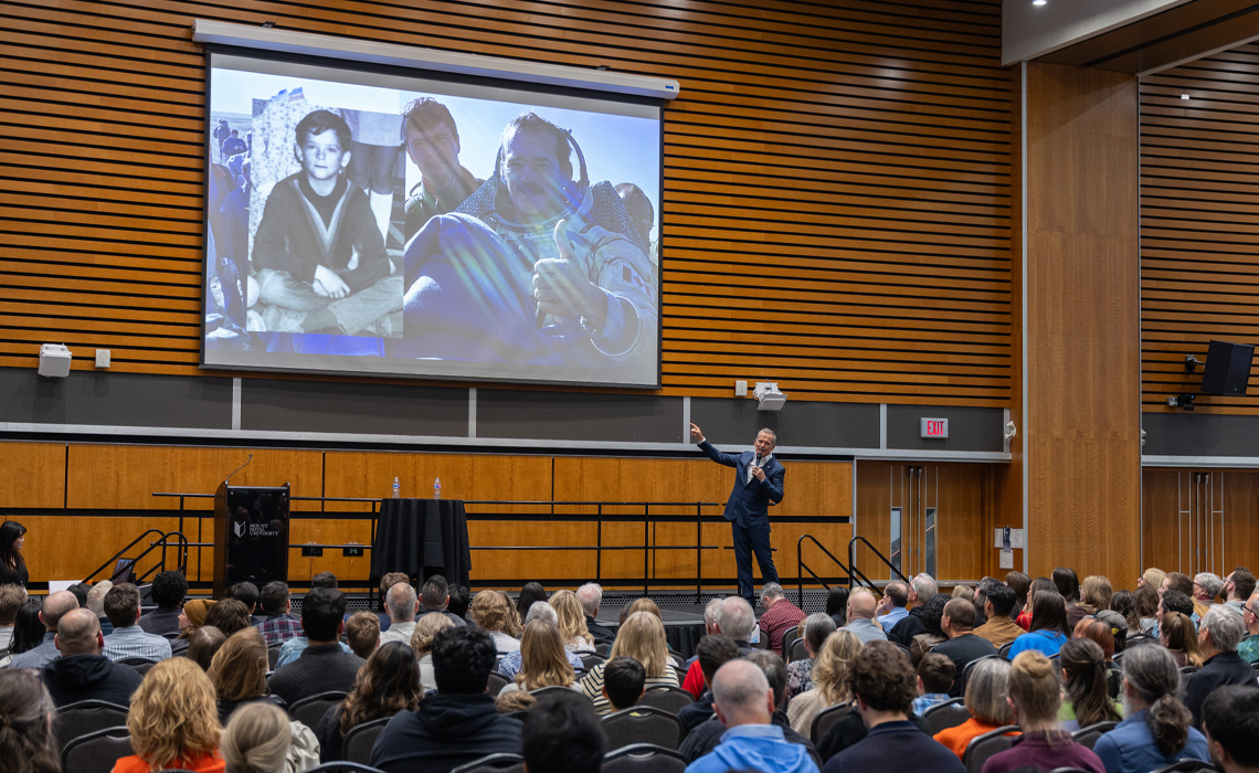 Colonel Chris Hadfield presenting to a large audience in a well-lit conference hall.