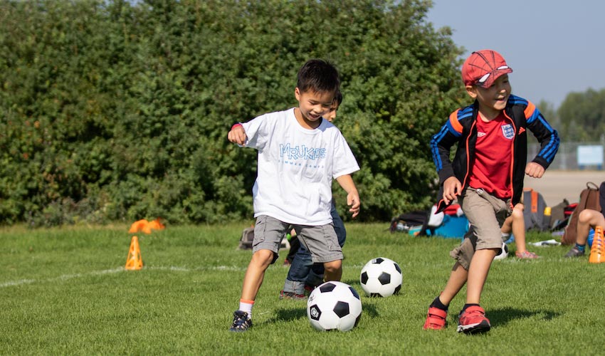 Several kids run around and kick soccer palls as a part of an MRUKids summer camp.