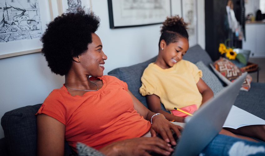 Happy mother smiling and working from home using a laptop while her young daughter sits next to her doing homework.