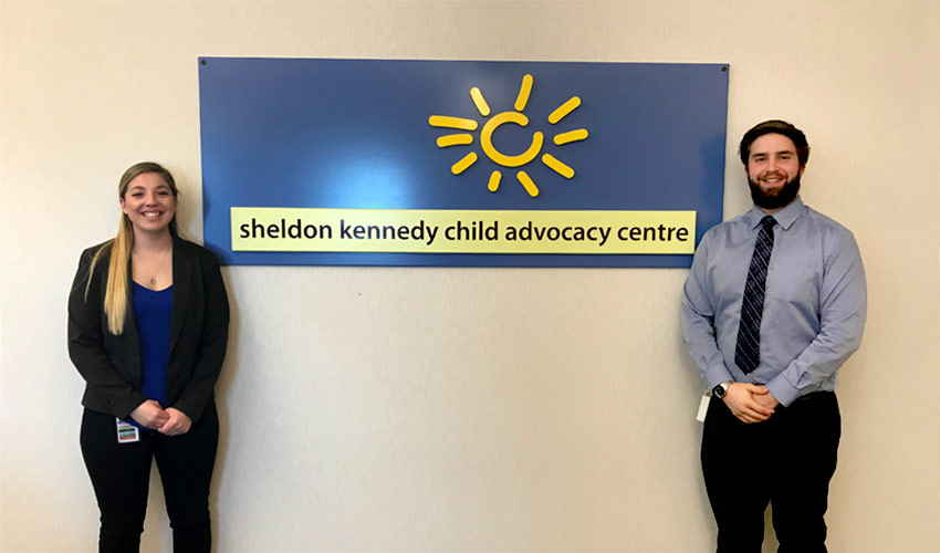 Nicole Erhardt and Benjamin Reid pose beside the sign for the Sheldon Kennedy Child Advocacy Centre