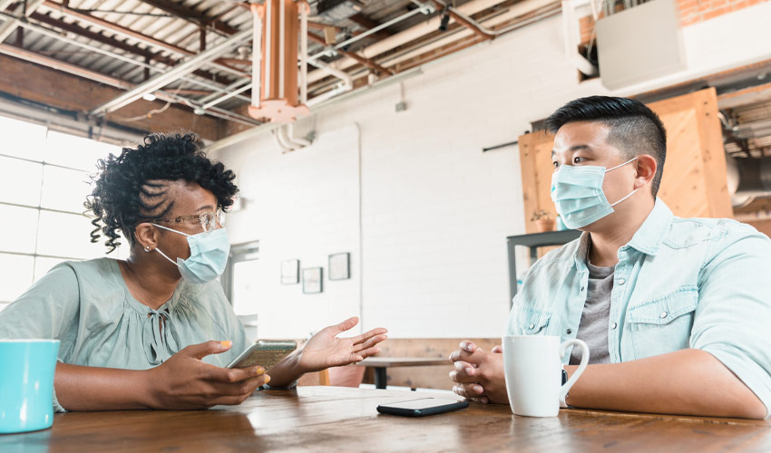 A businesswoman gestures as she meets with a male colleague over coffee. She is holding a smartphone. They are wearing protective face masks as they are meeting during the COVID-19 pandemic.
