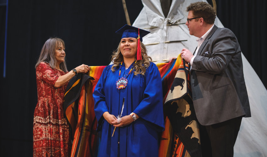 Lynsey Own Chief, who graduated with a Bachelor of Arts - Anthropology, is blanketed by linda manyguns, phd, associate vice-president, Indigenization and decolonization, left, and Chris Rogerson, executive director, student affairs.