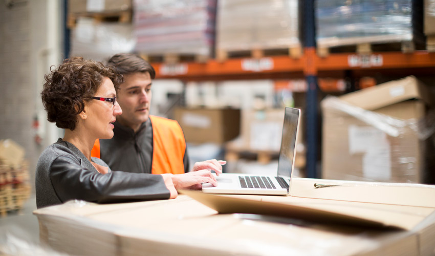 Two warehouse workers having a meeting, one is working on a laptop.