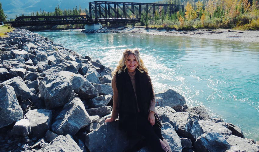 Megan Brawner sitting next to the Bow River with the Canmore Engine Bridge in the background.