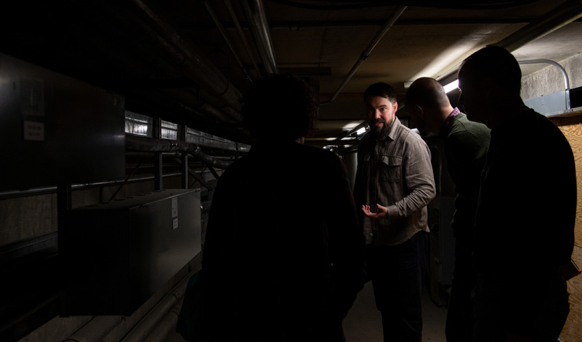 Manager of Building Operations Dylan Pritchard (centre), and Critical Systems Program Coordinator Glen Hogan (in silhouette, far right) lead a tour. As a 35-year employee, Hogan has spent more time in the tunnel than probably anyone else on campus.