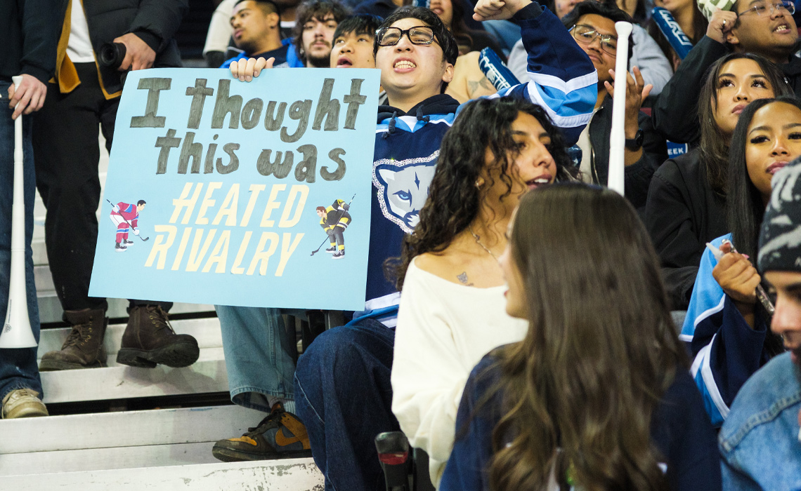 A hockey fan in the stands at the Crowchild Classic game, holding up a handmade sign that reads "I thought this was HEATED RIVALRY" in bold letters.
