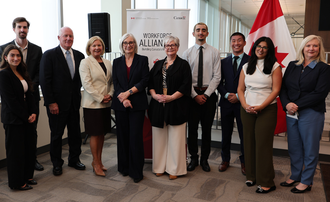 A group of people stands proudly in front of a large Canadian flag, smiling and posing for the photo.