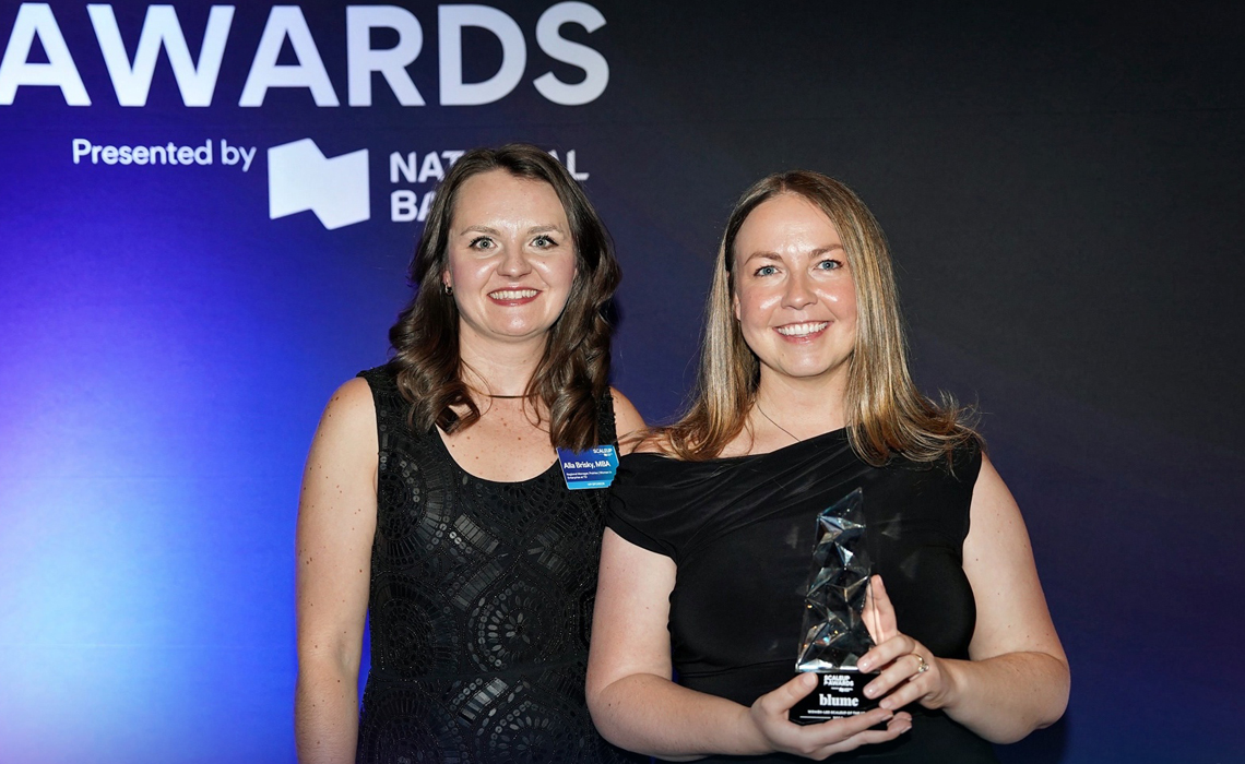 Two women proudly holding an award, standing in front of a blue wall.