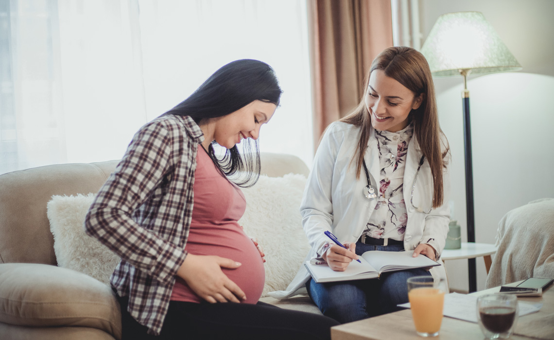 A pregnant woman sits on a couch while a midwife writes notes on a clipboard beside her.
