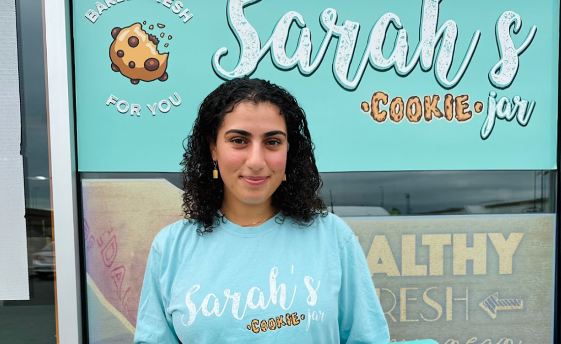 Sarah Omar poses in front of a sign that says "Sarah's Cookie Shop."