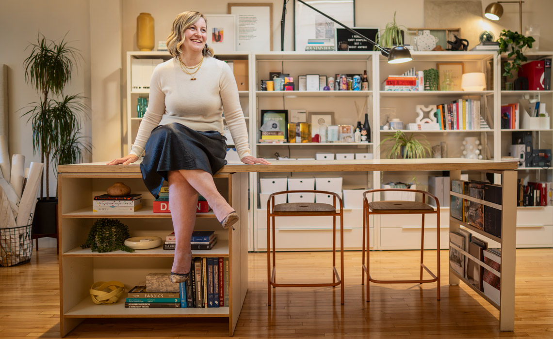 Sarah Ward sitting on a desk, surrounded by bookshelves filled with various books.