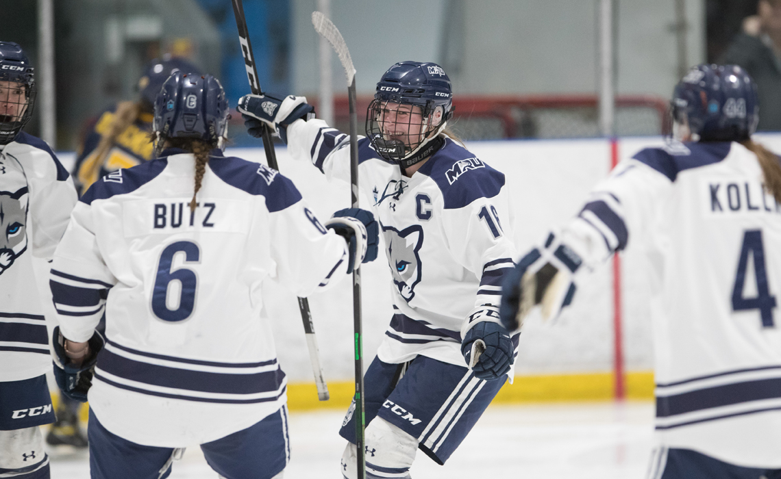 Tatum Amy and the women's hockey team joyfully celebrates scoring a goal, with players embracing and cheering on the ice.