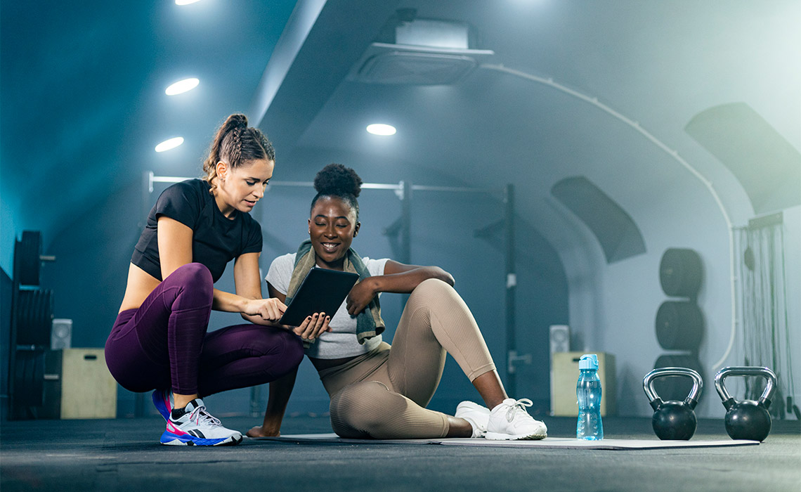 Two women in athletic wear review data on a digital tablet in a gym. Kettlebells and a water bottle are visible in the foreground.
