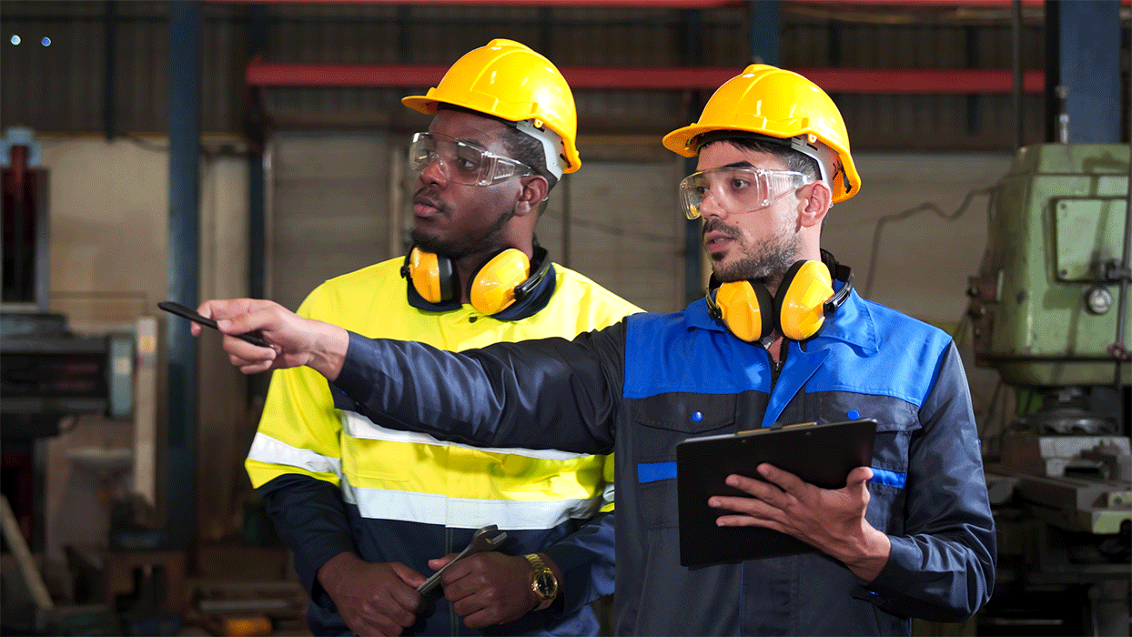 Two men in hard hats and safety glasses, gesturing towards an object while engaged in a conversation on-site.