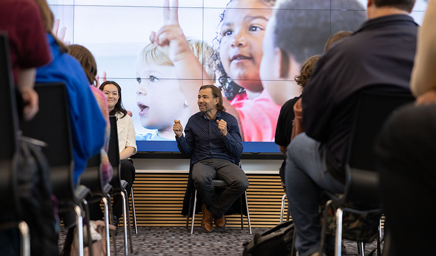 A man in front of a screen, talking excitedly in front rows of full seats.