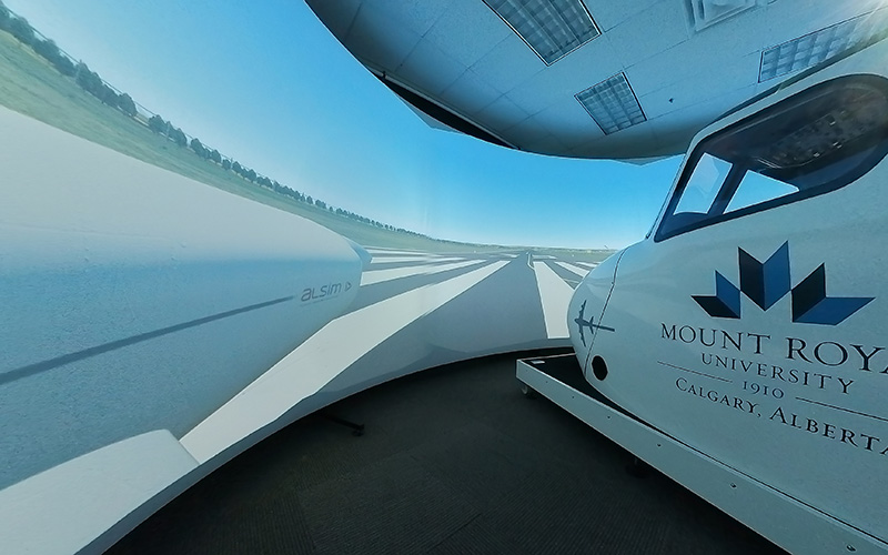 The nose of an airplane in a dark room in front of a projection of a runway.