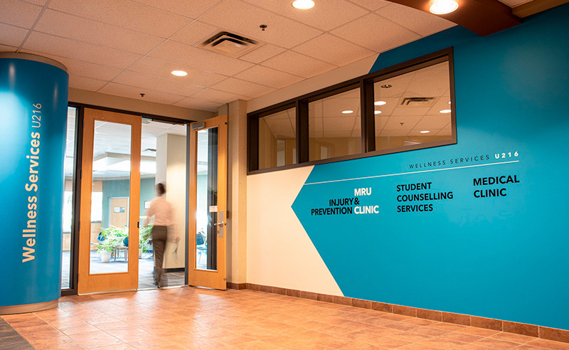 The entrance to MRU Wellness Services, which is a bright blue wall and blue pillar with department names in vinyl and a person walking into the door.