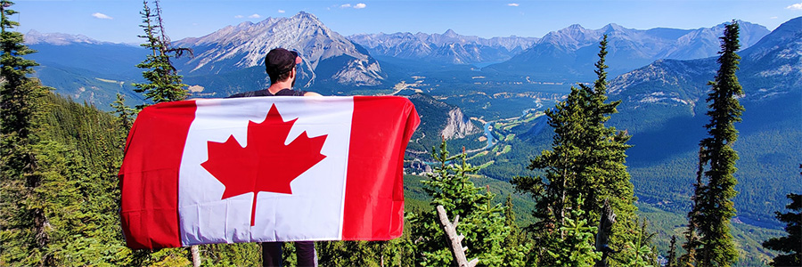 A person holding the Canadian flag like a cape while looking down on the town of Banff from a mountaintop.