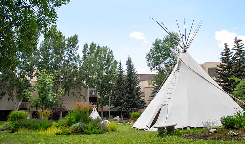 Photo of the Centennial Garden. Small garden sections of shrubs and grasses frame a full-sized and small teepee in front of a brick and concrete building.