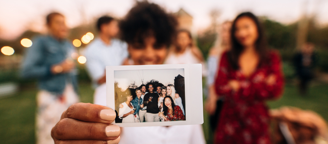 A person holding up a polaroid photo to the camera at a social gathering.
