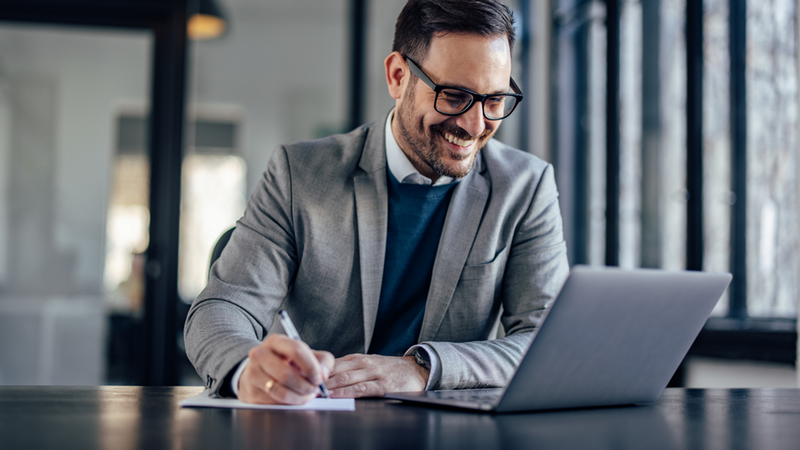 A man at a desk with a laptop happily writing notes