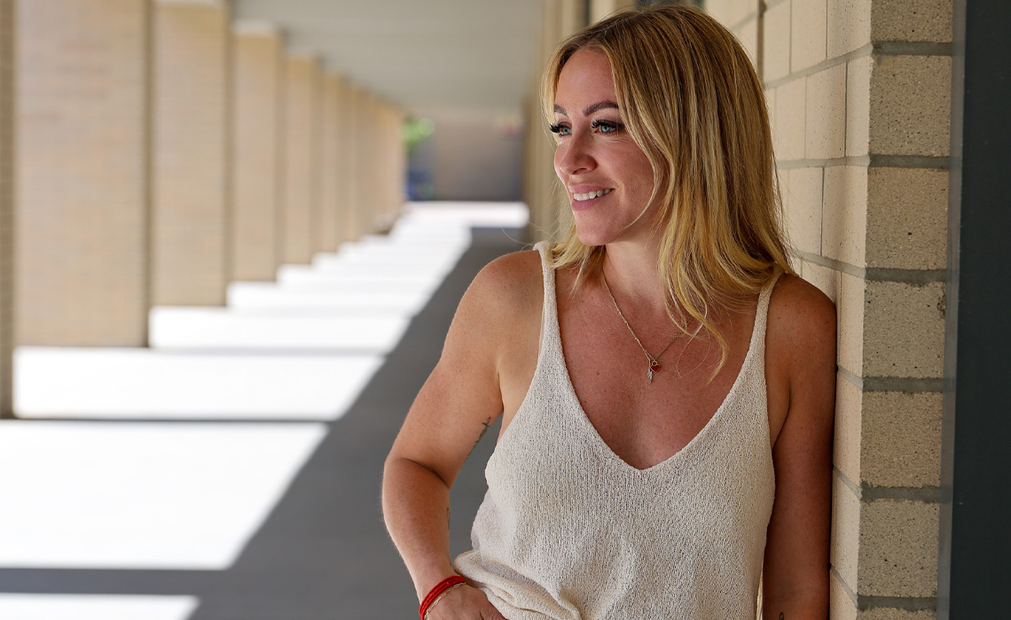 Amanda Hamilton in jeans and a white tank top leans casually against a wall outside of the EA building at MRU.
