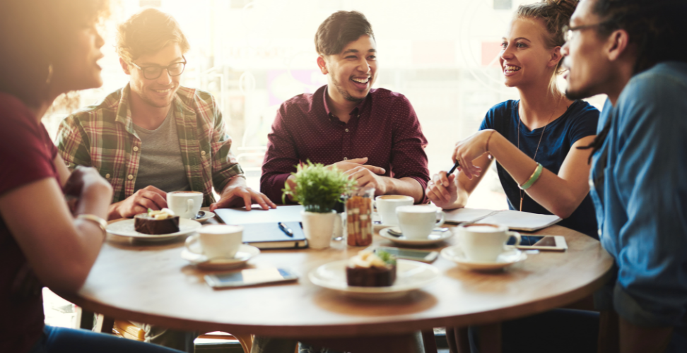 A group of people at a large round table in chatting with snacks and coffees.