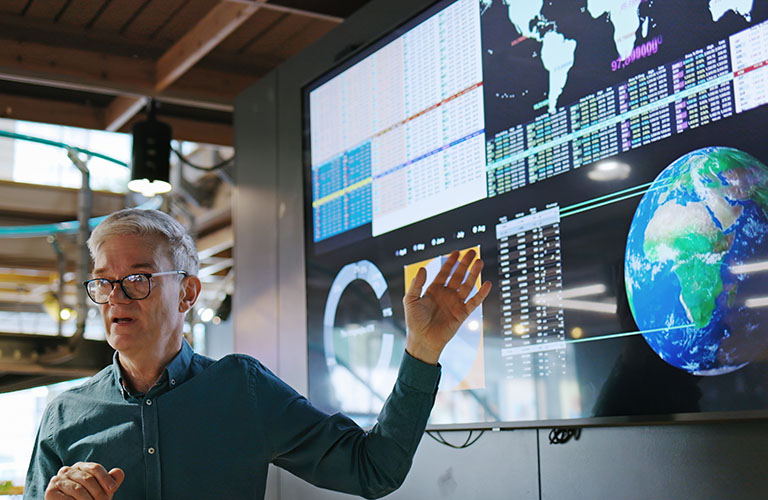 A man presenting in front of a screen displaying graphs, data and an image of the earth.