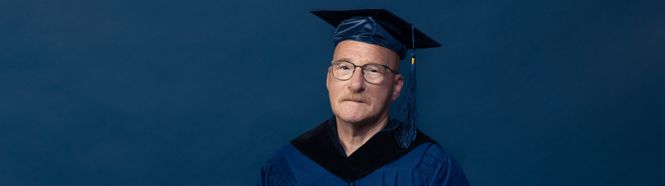 A man with glasses in a graduation cap and gown, with a blue background.