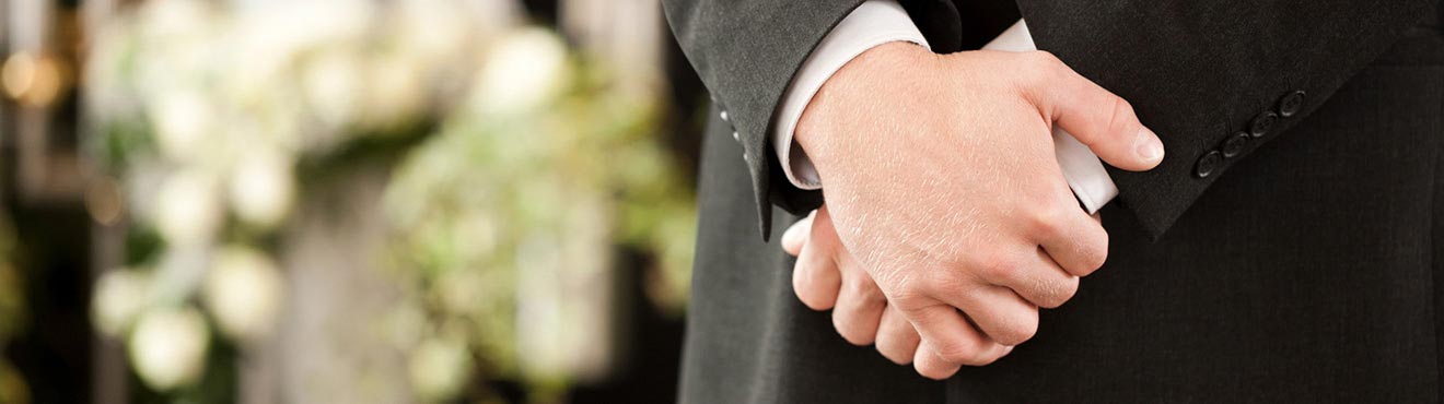 Politely clasped hands of a funeral director in front of some funeral bouquets.