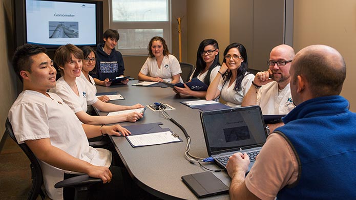 A group of massage therapy students around a large table listening to an instructor give a presentation.