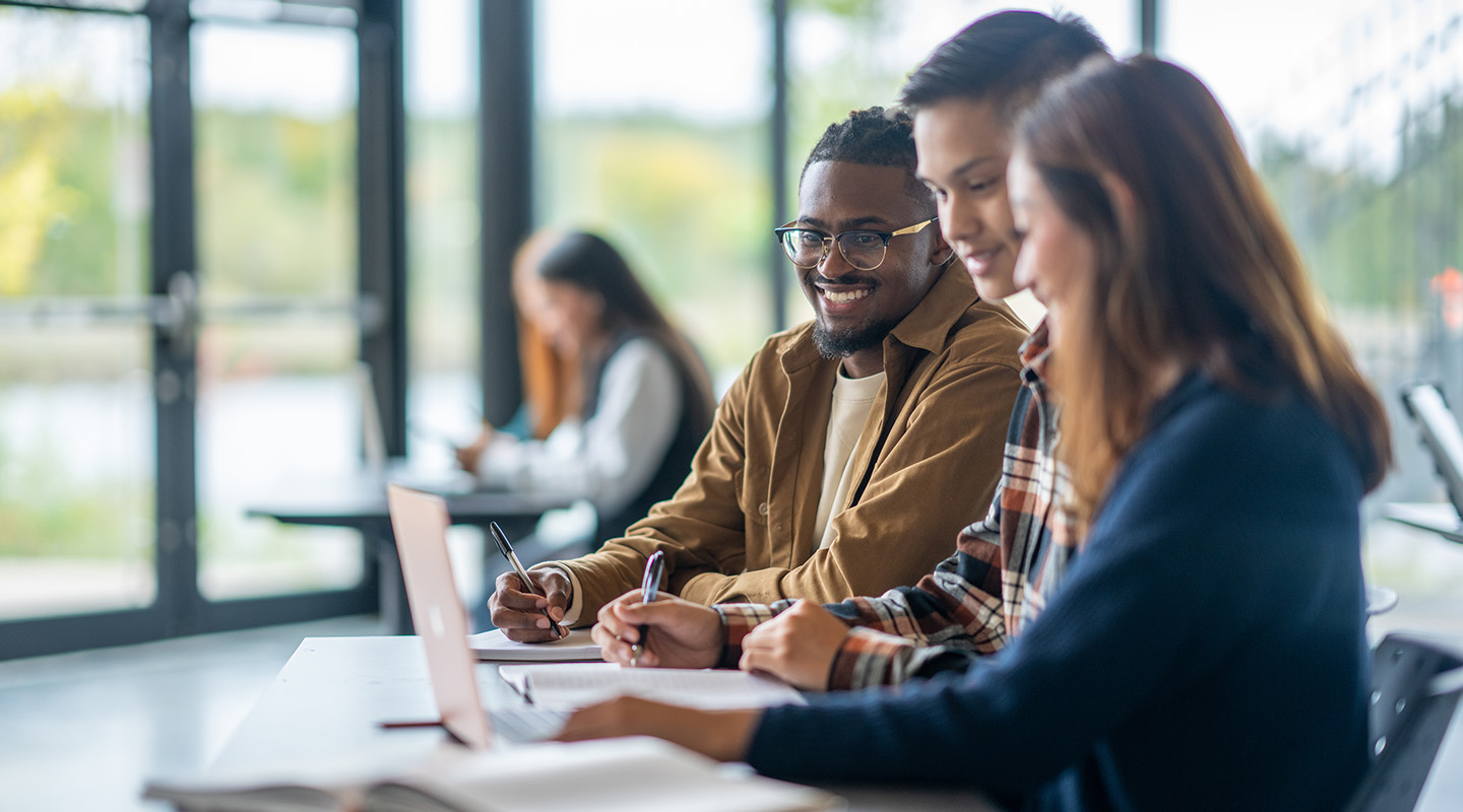 Three students are sitting together in at a desk, taking notes and chatting beside an open laptop.