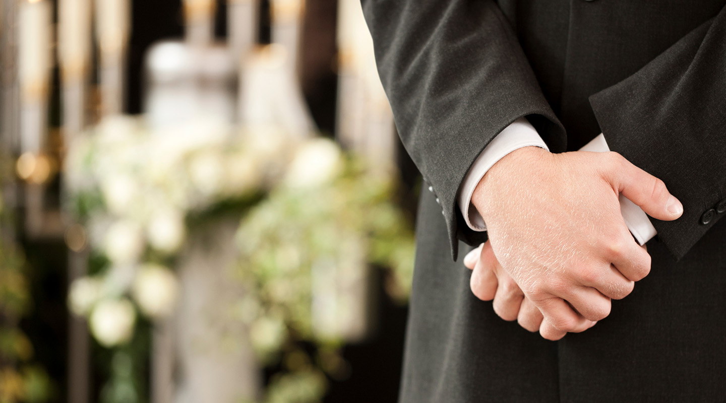 Politely clasped hands of a funeral director in front of some funeral bouquets.