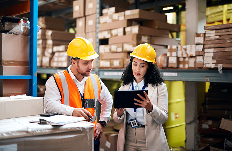 Two people wearing safety vests and hardhats in a warehouse reviewing a tablet.