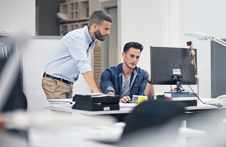 A professional working on a computer while a coworker reviews their work.