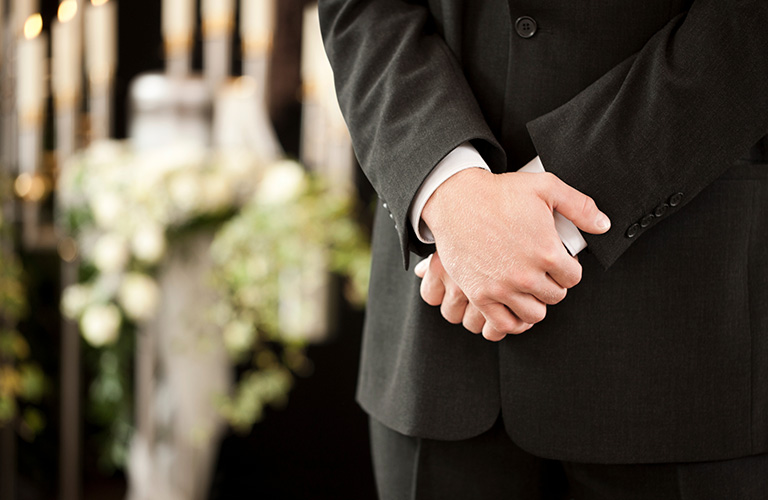 Politely clasped hands of a funeral director in front of some funeral bouquets.