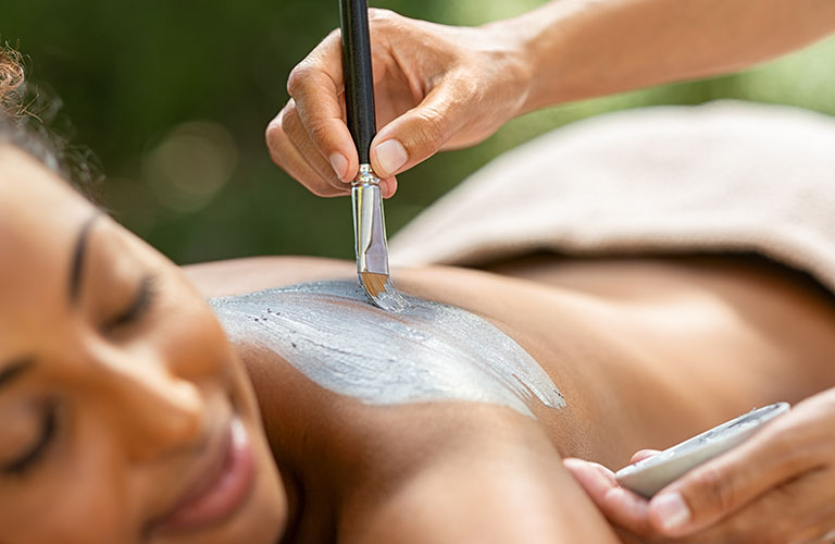 Hands using a brush to spread mud on the back of a spa patient.