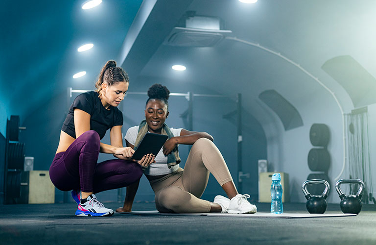 Fitness gym instructor showing workout plan on tablet to their client. They are sitting on the gym floor, resting after workout.