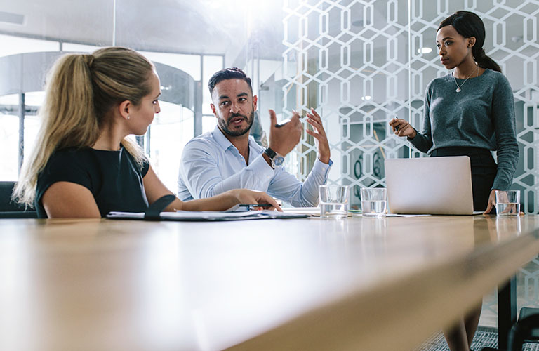 A business woman stands at a table listening to two people having a conversation.